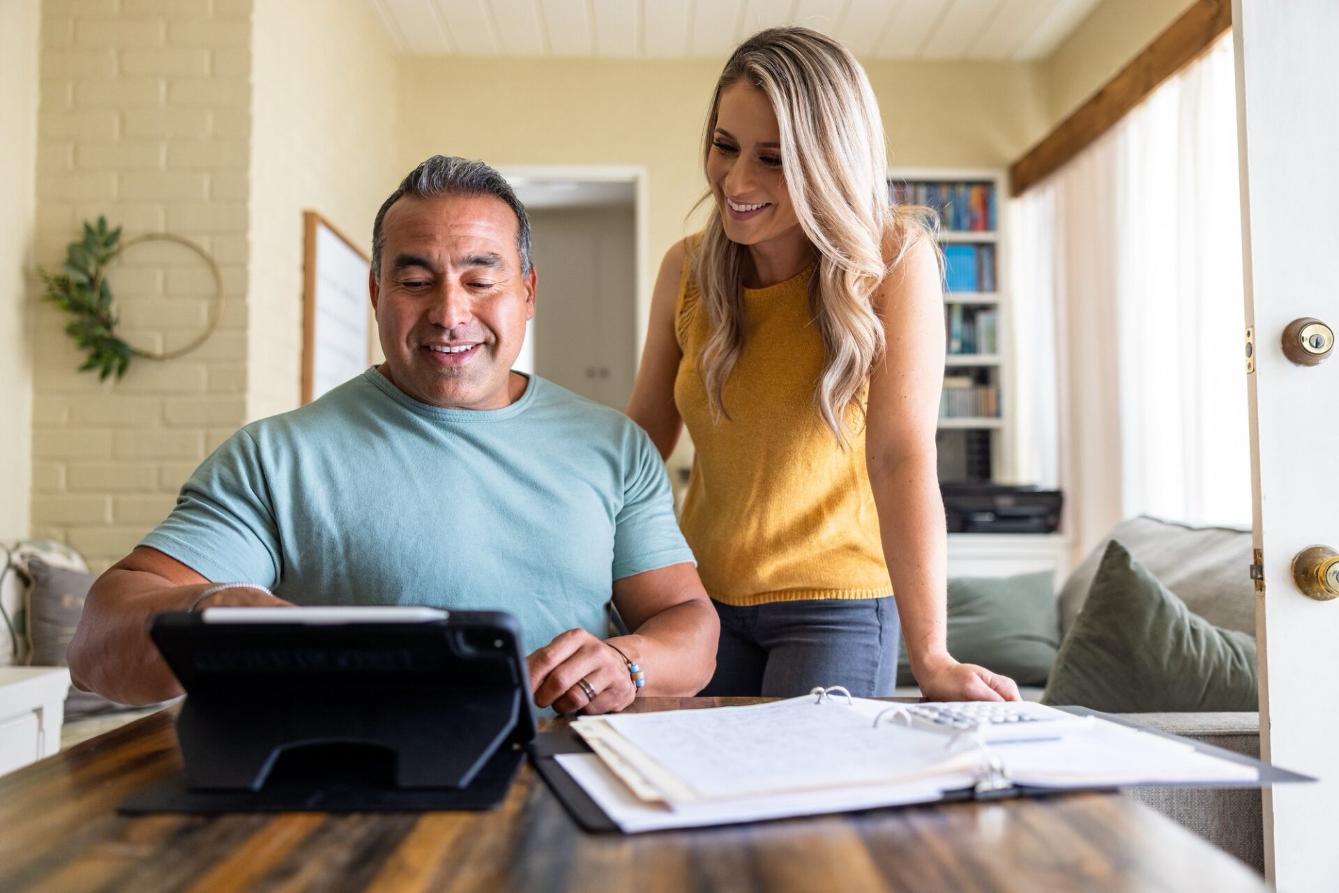 A man and woman smiling while reviewing documents and using a tablet at a kitchen table.