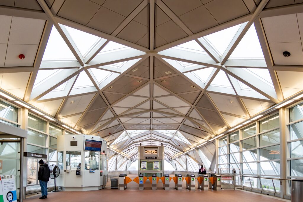 Interior view of a modern train station entrance with a geometric glass ceiling, ticket barriers, and an information booth.