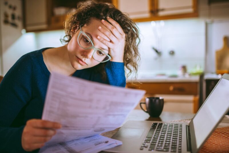 A woman in glasses reviewing tax documents with a laptop at home.