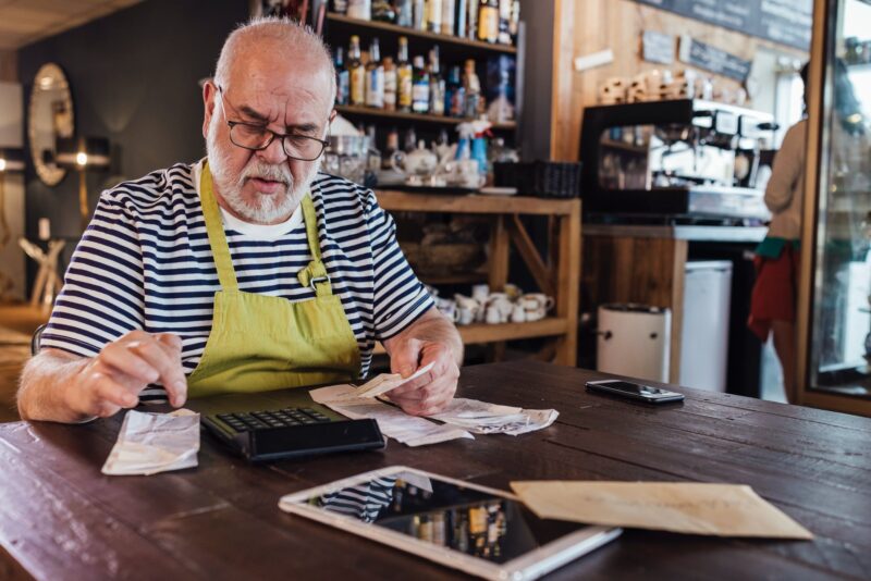 An elderly man wearing an apron, calculating expenses at a small business.